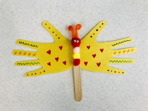 A yellow butterfly made out of two hand-shaped pieces of paper for the wings and a popsicle stick for the body.